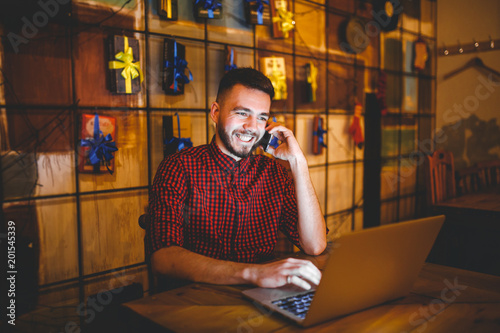 Young handsome Caucasian man with beard and toothy smile in red shirt works behind laptop, hands on keyboard sitting at wooden table. Uses calls on mobile phone. In evening at the coffee shop