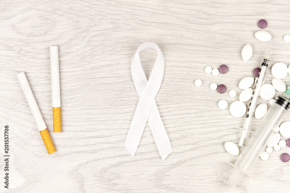 Oncological disease concept. White ribbon as a symbol of lung cancer, two сigarettes, pills and syringes isolated on light wooden background Oncological disease concept. White ribbon as a symbol of lung cancer, two сigarettes, pills and syringes isolated on light wooden background