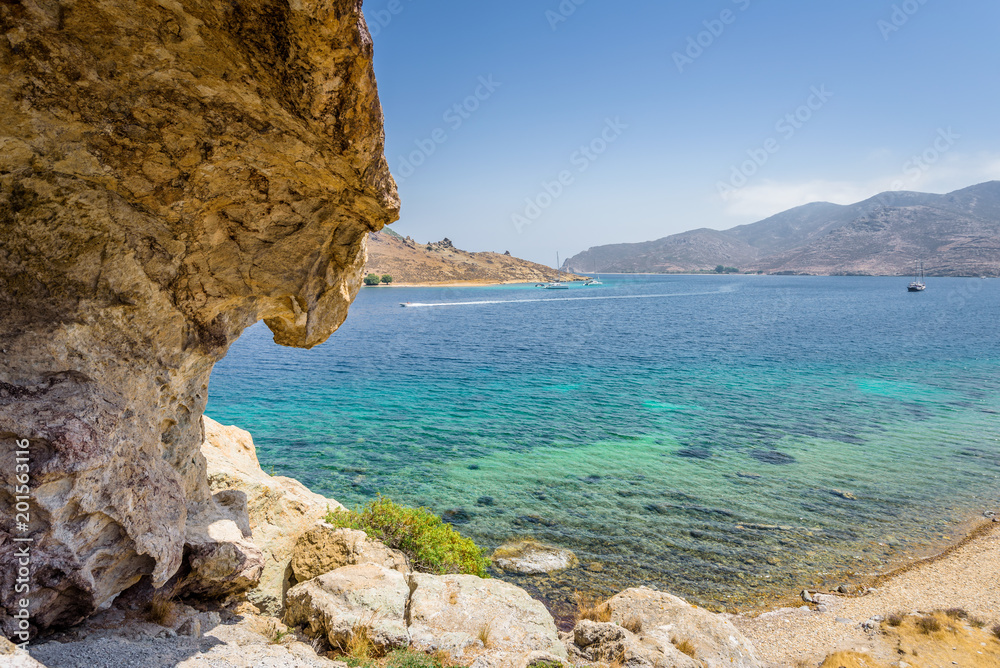 Fototapeta premium Beautiful sunny view through a rocky cave to the greek blue sea with crystal clear water from an a hill with boats cruising fishing surrounded by mountains, Patmos Island, Kos, Dodecanese/ Greece