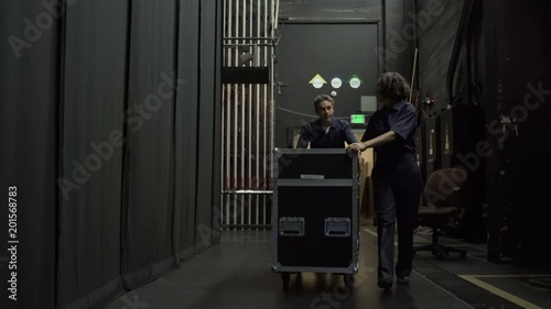 Two stage workers wheeling equipment trolley towards camera, backstage photo