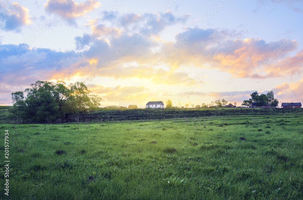 Empty green meadow on a background of country house and rising sun.