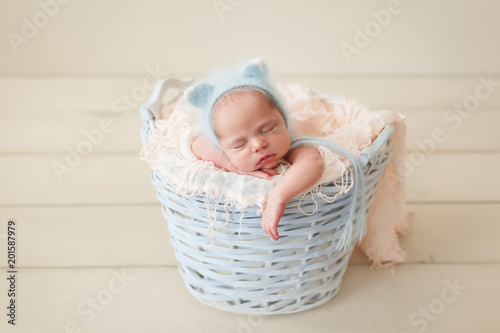 Newborn sleeping in a wicker basket in a blue fluffy knit kitty hat on a pink props on a beige wooden floor