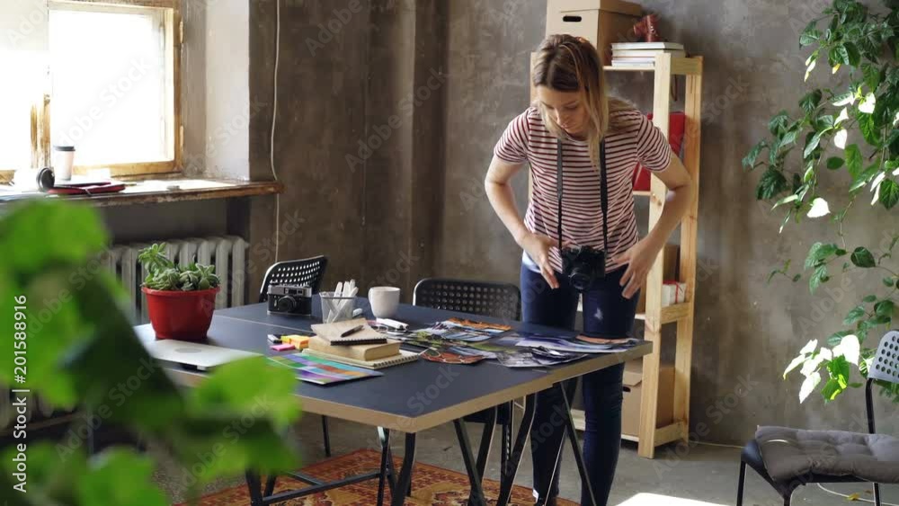 Young creative girl is making flat lay out of colorful photos, then shooting them with camera. Woman is standing near modern table and wearing casual clothes. Young creative girl is making flat lay out of colorful photos, then shooting them with camera. Woman is standing near modern table and wearing casual clothes.