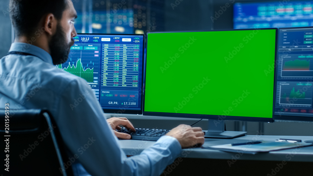 Over the Shoulder View of Stock Market Trader Working on a Computer with Isolated Mock-up Green Screen and Second Display Showing Number Ticker with Graphs. Over the Shoulder View of Stock Market Trader Working on a Computer with Isolated Mock-up Green Screen and Second Display Showing Number Ticker with Graphs.