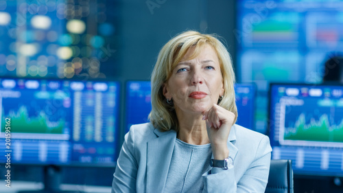 Stock Market Leading Analyst Thinking Hard on Solving Financial Problem. Behind Her People Working and Monitors Show Graphs and Figures.