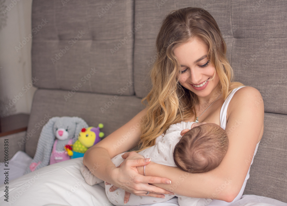 Breastfeeding baby. Pretty mother holding her newborn child. Mom smile and nursing infant. Beautiful woman and new born love at home. Blondе mother breast feeding baby.