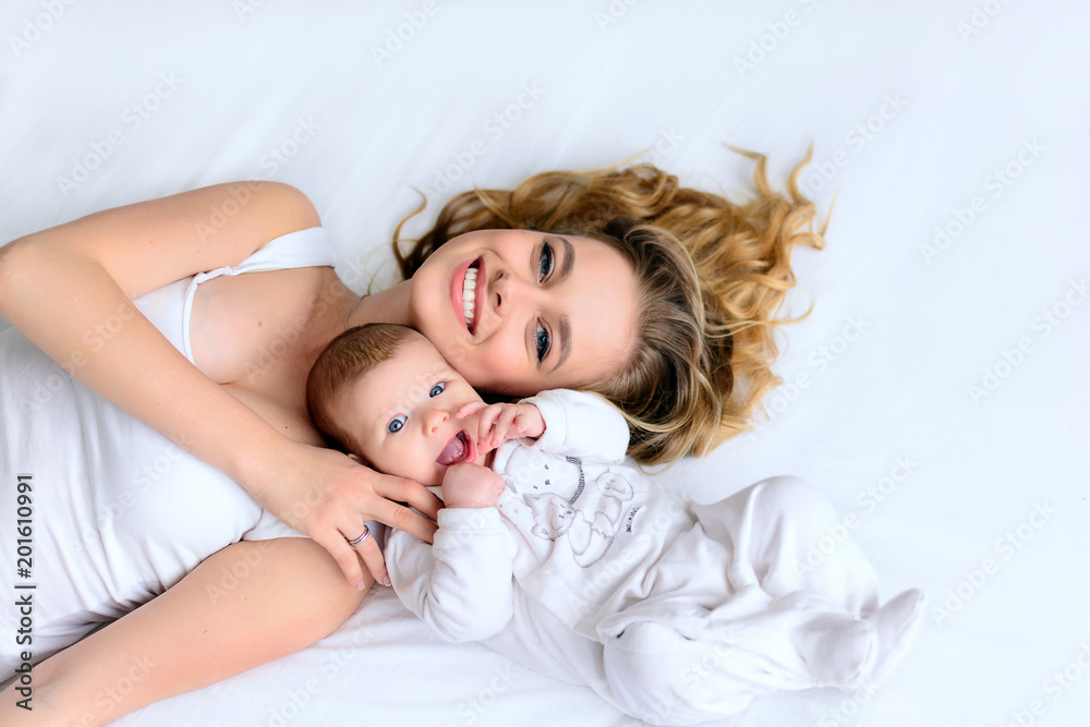 Beautiful mother and newborn baby lying in bed, hugging with love, smiling. Pretty blonde woman and happy infant lay together on white backgound of bed. From high angle shot, above view.
