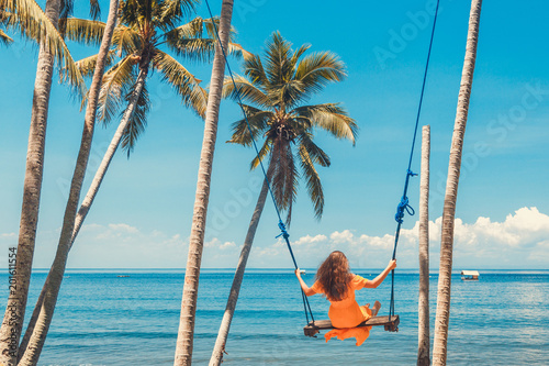 Vacation concept. Happy young woman in yellow dress sitting on swing enjoying sea view.