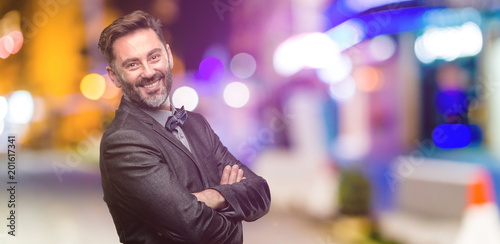 Middle age man, with beard and bow tie with crossed arms confident and happy with a big natural smile laughing at night club