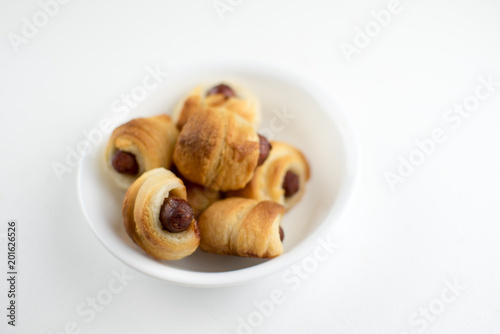 Home made pigs in a blanket. Sausages rolled in croissant dough baked and placed in a white serving dish. On white background.