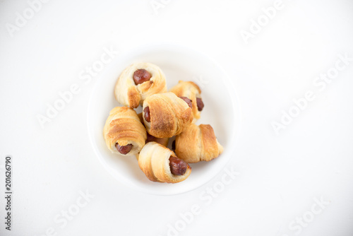 Home made pigs in a blanket. Sausages rolled in croissant dough baked and placed in a white serving dish. On white background.