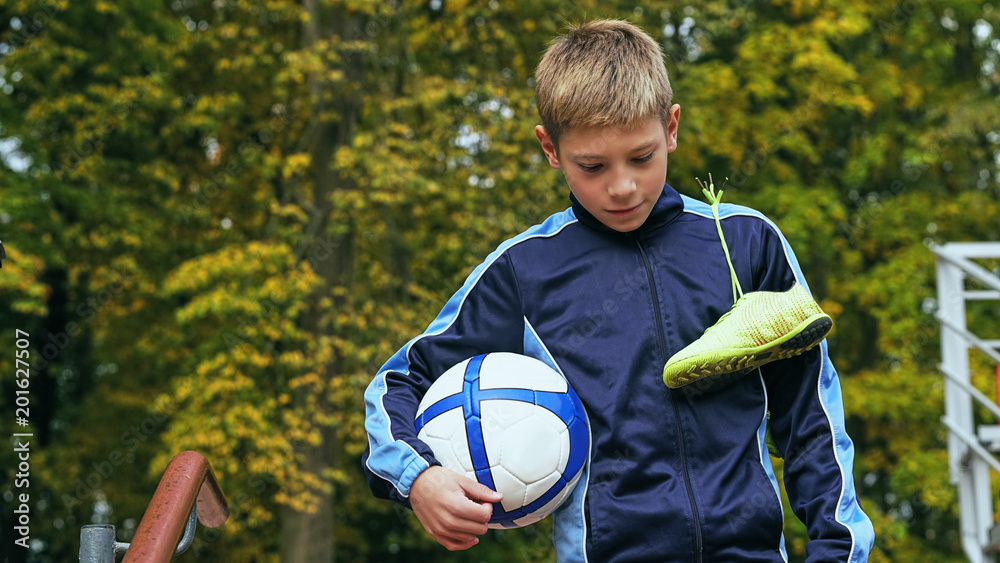 Obraz premium Smiling teenage boy with a soccer ball in his hand and soccer boots on the shoulder against the background of the stadium. Sports training in the field.