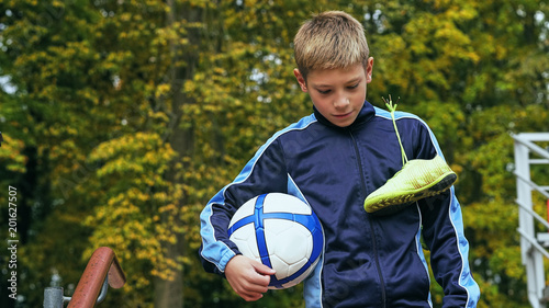 Smiling teenage boy with a soccer ball in his hand and soccer boots on the shoulder against the background of the stadium. Sports training in the field.