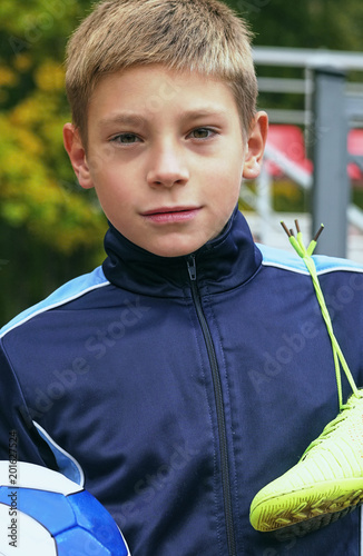 Smiling teenage boy with a soccer ball in his hand and soccer boots on the shoulder against the background of the stadium. Sports training in the field.