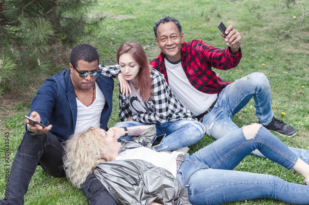 group of smiling friends with smartphones sitting on grass in park