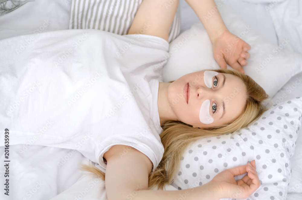 Under Eye Treatment. Woman Face With Patches Under Eyes. Portrait Of Gorgeous Smiling Female In Sleepwear Resting On White Bedding In Light Bedroom Under Eye Treatment. Woman Face With Patches Under Eyes. Portrait Of Gorgeous Smiling Female In Sleepwear Resting On White Bedding In Light Bedroom