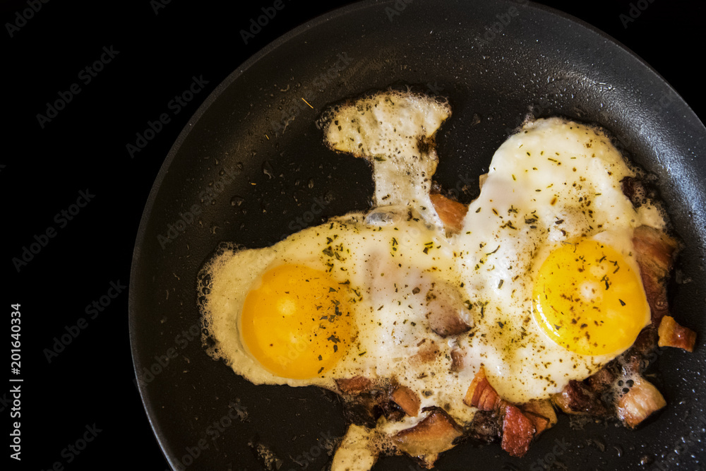 fried eggs and bacon in a frying pan on black background