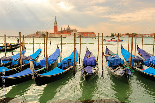 Anchored gondolas standing in a row between piles against San Giorgio Madjore. Sunlight through the clouds. Venice