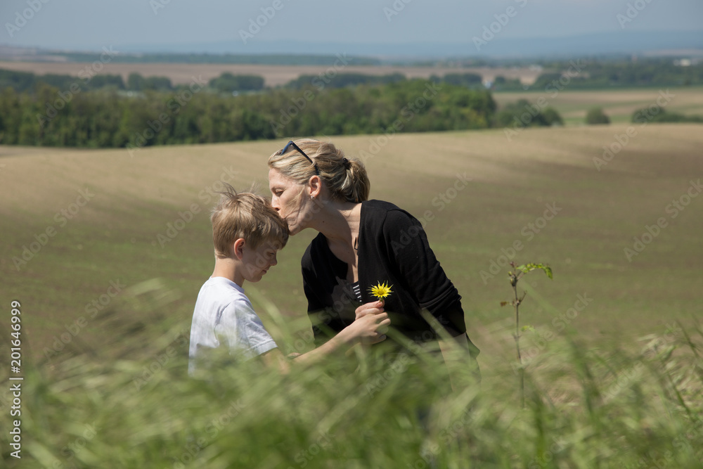 Young blonde boy gives flower to his mother with love. Outdoors, natural scene.