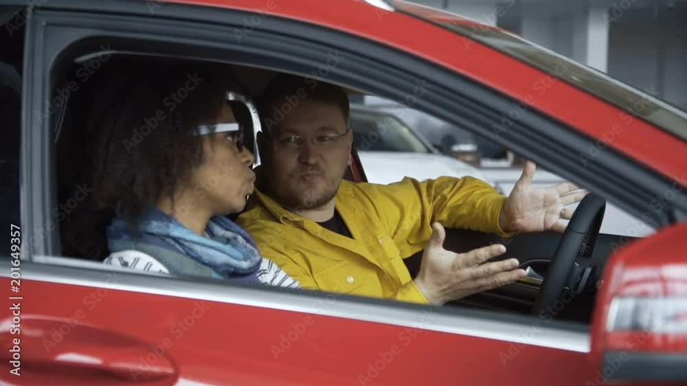 Side view of content multiethnic man and woman sitting inside of car in auto shop and discussing purchase. Side view of content multiethnic man and woman sitting inside of car in auto shop and discussing purchase.