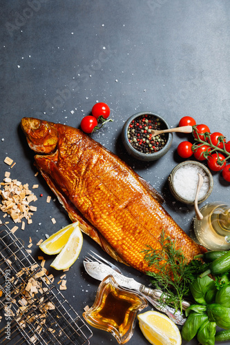 Smoked whitefish with salt and pepper in dish, fork and knife, lemon, basil, tomatoes, little cucumbers, parslay, grid for grilling, cherry wood chunks  on a black background, top view, overhead, flat photo