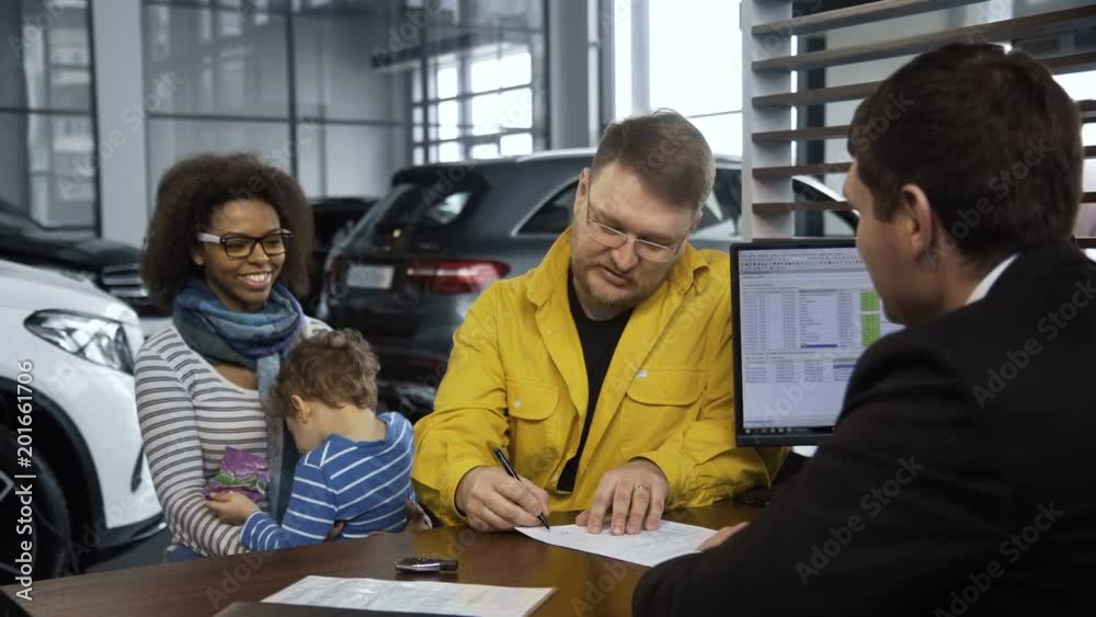 Happy multiethnic couple with son making purchase of car sitting at table with dealer and signing papers.