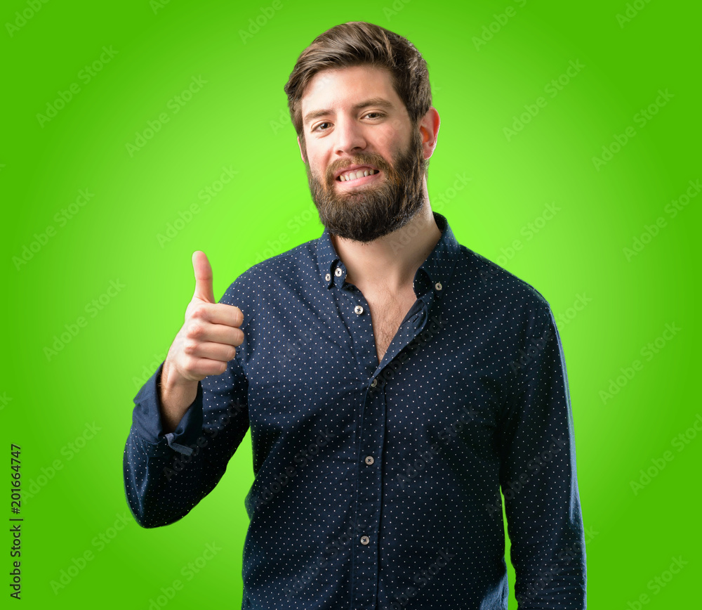 Young hipster man with big beard smiling broadly showing thumbs up gesture to camera, expression of like and approval over green background