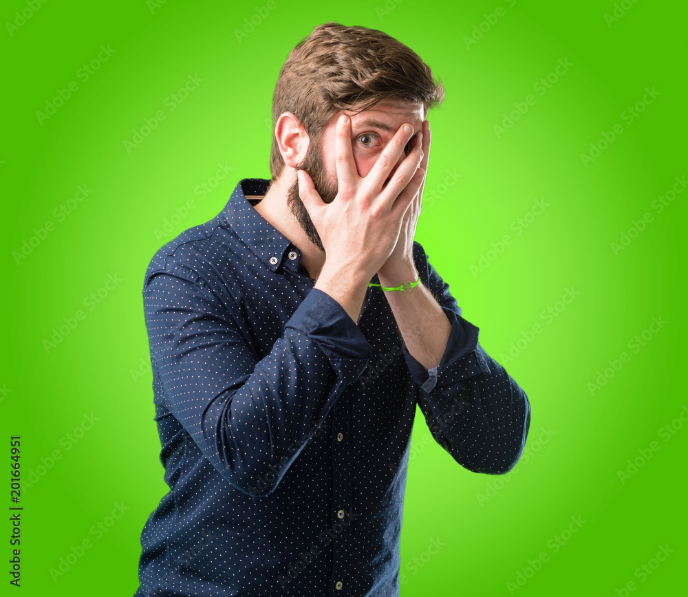 Young hipster man with big beard smiling having shy look peeking through her fingers, covering face with hands looking confusedly broadly over green background Young hipster man with big beard smiling having shy look peeking through her fingers, covering face with hands looking confusedly broadly over green background
