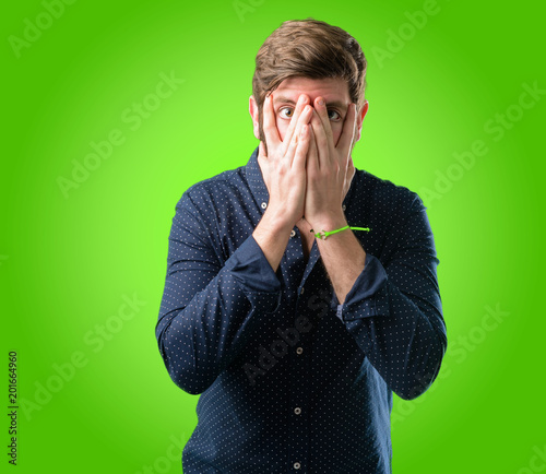 Young hipster man with big beard smiling having shy look peeking through her fingers, covering face with hands looking confusedly broadly over green background