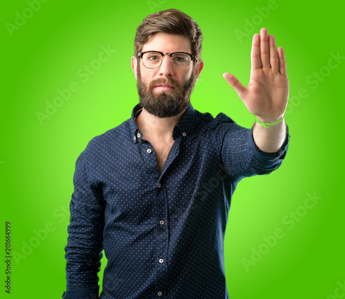 Young hipster man with big beard annoyed with bad attitude making stop sign with hand, saying no, expressing security, defense or restriction, maybe pushing over green background