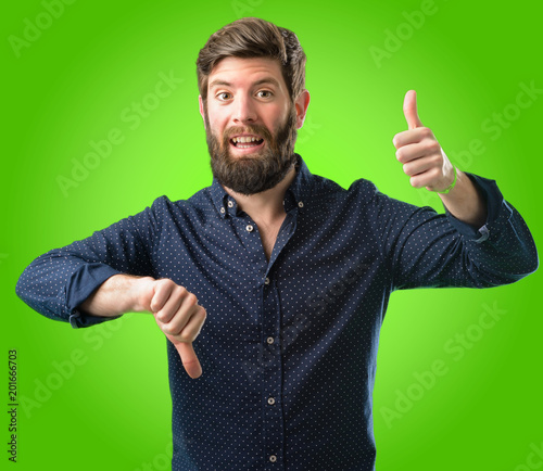 Young hipster man with big beard confused with thumbs up and down, trying to take a decision expressing doubt and frustration over green background