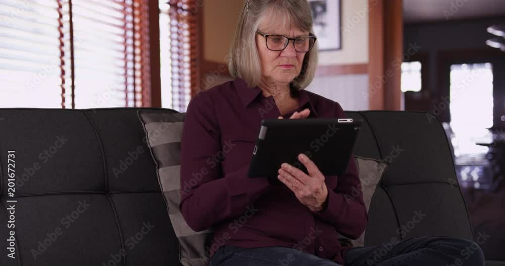 Lively mature woman using portable tablet to browse online in living room, White senior lady relaxed on sofa typing on handheld pad device inside her home, 4k Lively mature woman using portable tablet to browse online in living room, White senior lady relaxed on sofa typing on handheld pad device inside her home, 4k