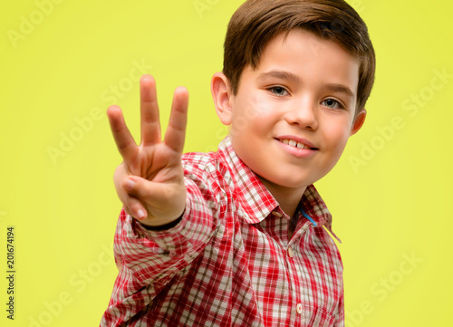 Handsome toddler child with green eyes raising finger, is the number three over yellow background