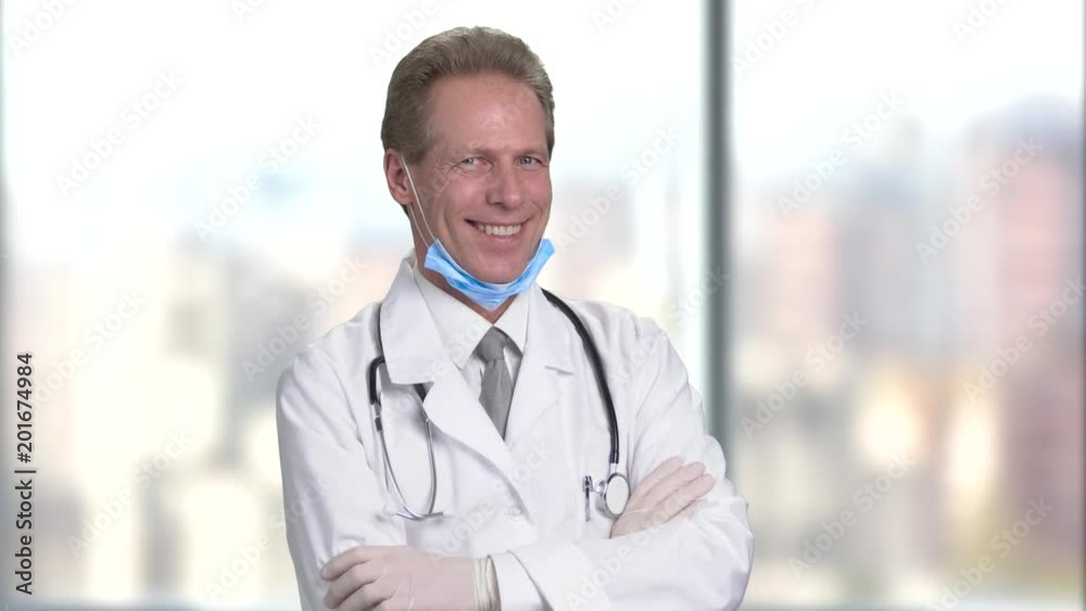 Happy mature middle aged smiling doctor with folded arms, front view. Bright abstract blurred windows background with view on city. Happy mature middle aged smiling doctor with folded arms, front view. Bright abstract blurred windows background with view on city.