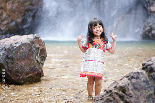 Asian children cute or kid girl smile with fun and enjoy playing with show two finger for take a photo on waterfall with stone or rock and holiday relax trip for summer at Jokkradin waterfall