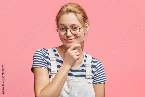 Lovely blonde female model holds chin, looks with cunning and delighted expression, has intriguing idea in mind, wears stripes t shirt and denim overalls, poses against pink background alone