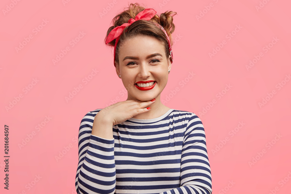 Candid shot of pleasant looking female with make up, has positive expression, keeps hand under chin, wears striped sweater and stylish headband, poses against pink background. Happiness and people Candid shot of pleasant looking female with make up, has positive expression, keeps hand under chin, wears striped sweater and stylish headband, poses against pink background. Happiness and people