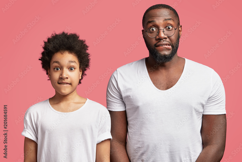 Horizontal shot of attractive dark skinned man and small male child look in bewilderment and shocked expressions, bite lower lips, admire something with delighted look, isolated over pink wall Horizontal shot of attractive dark skinned man and small male child look in bewilderment and shocked expressions, bite lower lips, admire something with delighted look, isolated over pink wall