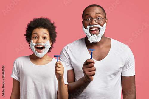 Photo of happy schoolboy with Afro hairdo attempts to shave like his dad, applies shaving gel on face and takes razor, wants to become older, stands next to father, enjoy free time together.