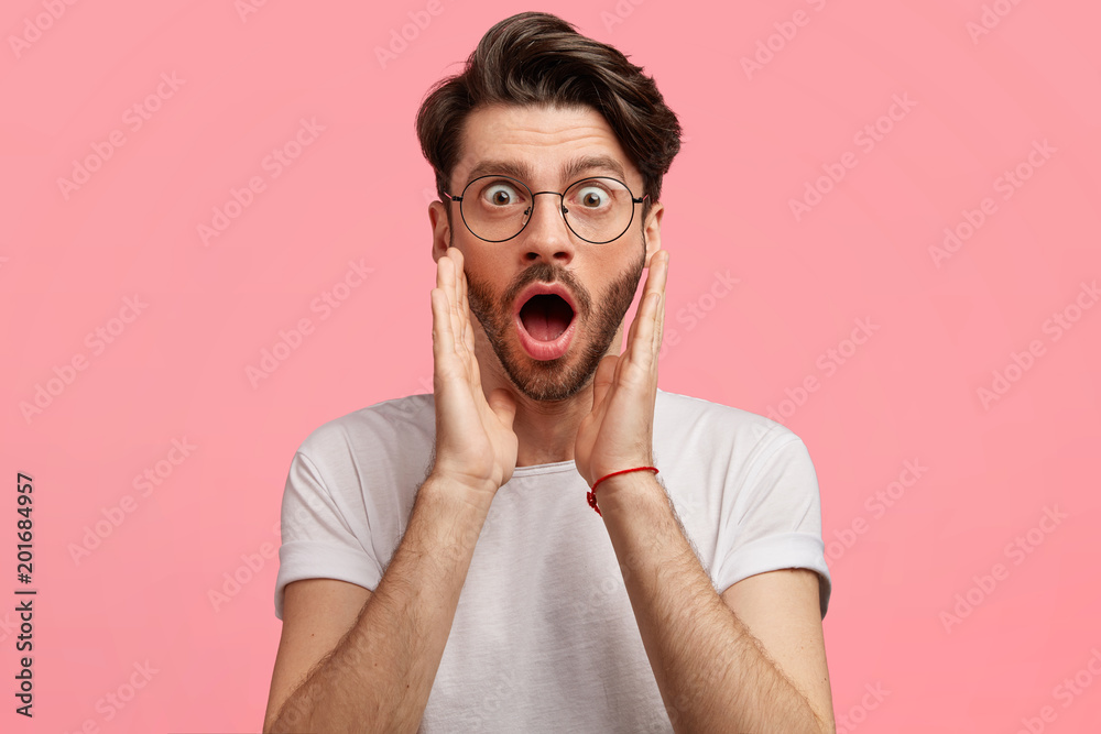 Handsome stylish bearded young male with stylish hairdo, looks with shocked amazed expression, expresses wonder after failing exam, dressed in casual white t shirt. Fashionable guy poses indoor Handsome stylish bearded young male with stylish hairdo, looks with shocked amazed expression, expresses wonder after failing exam, dressed in casual white t shirt. Fashionable guy poses indoor