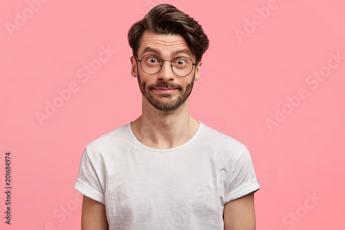 Portrait of attractive young male model has shocked pleased expression, trendy hairdo, wears round spectacles, hears something unexpected. Stunned student surprised to pass exam successfully