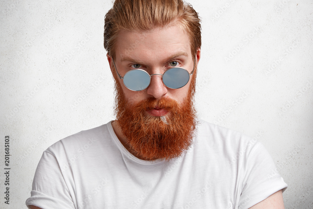 Confident male shop assistant advertises new fashionable shades as new trend this summer, has attractive appearance, dressed in casual white t shirt, isolated over concrete wall. People and style Confident male shop assistant advertises new fashionable shades as new trend this summer, has attractive appearance, dressed in casual white t shirt, isolated over concrete wall. People and style