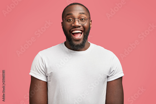 Waist up portrait of glad dark skinned handsome male model with happy expression, wears round spectacles, being in good mood as recieve bonus for diligent work, isolated over pink background