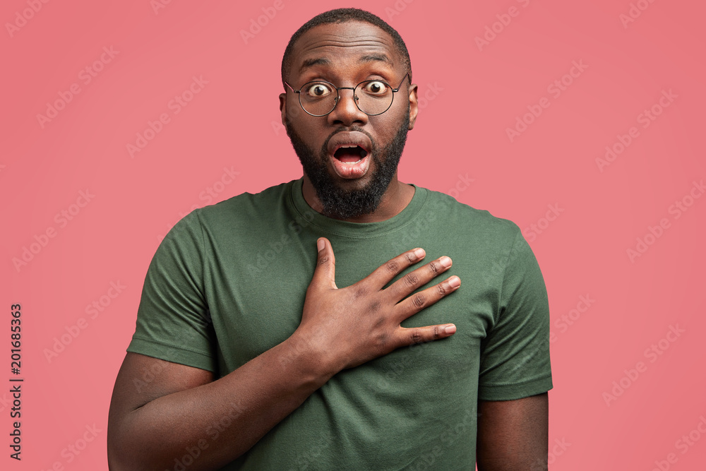 Surprised African American man keeps hands on chest, jaw dropped out, hears  shocking news, isolated over pink background. Emotional dark skinned young  male wears casual t shirt and spectacles Stock Photo
