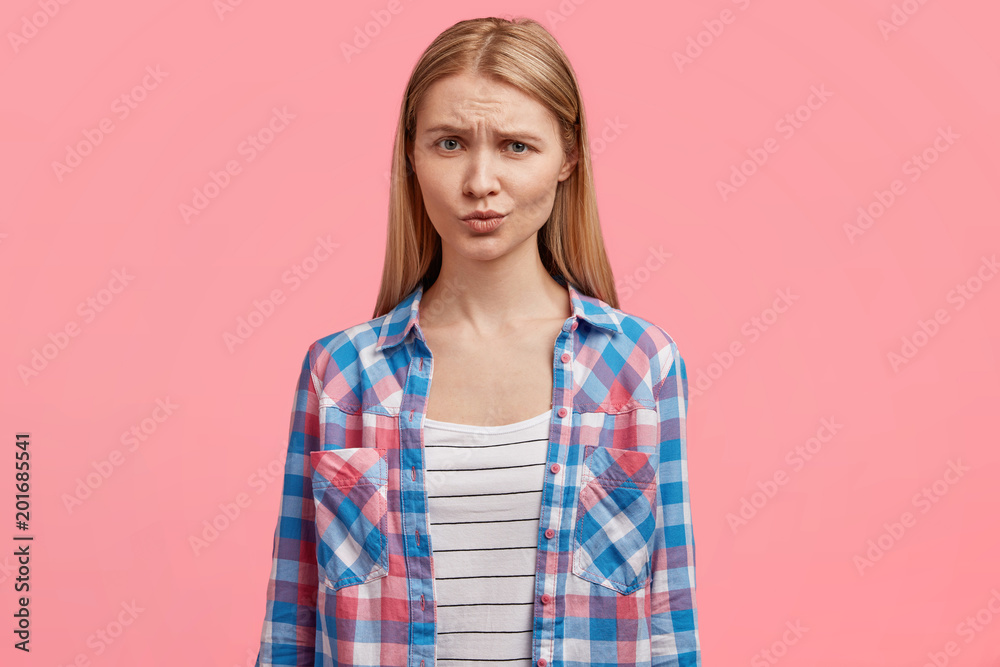 Waist up portrait of pleasant looking blonde female with healthy skin, frowns face, feels insult after quarrel, dressed in casual checkered t shirt, has no make up, demonstrates natural beauty Waist up portrait of pleasant looking blonde female with healthy skin, frowns face, feels insult after quarrel, dressed in casual checkered t shirt, has no make up, demonstrates natural beauty