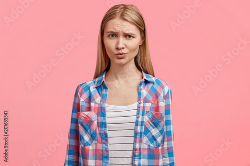 Waist up portrait of pleasant looking blonde female with healthy skin, frowns face, feels insult after quarrel, dressed in casual checkered t shirt, has no make up, demonstrates natural beauty