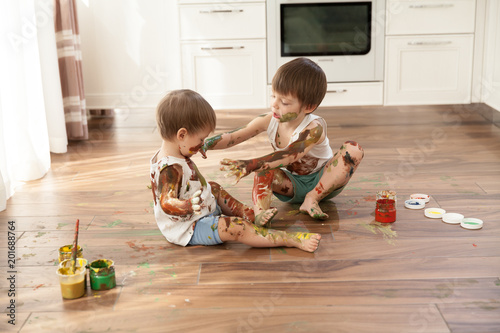  Two boys, two brothers playfully paint each other sitting on the floor.