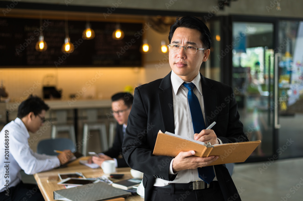 Asian businessman holding a book and pen to write a business plan. He standing and looking forward, thinking for business future and financial success.