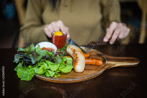Blurred hand of Woman holding knife and fork for eating food, close up Sausage with Tomato Sauce and green vegetable on wood plate