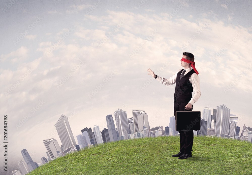 Young blindfolded businessman steps on a a patch of grass with a grey buildings in the background Young blindfolded businessman steps on a a patch of grass with a grey buildings in the background